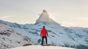 A person in a red jacket stands on a snowy peak, facing the Matterhorn mountain under a cloudy sky, embodying the spirit of "I Can Do All Things through Christ who Strengthens Me.