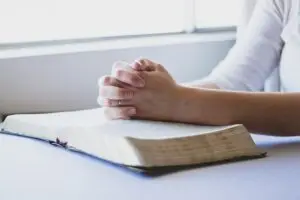 Hands clasped in prayer rest on an open book, near a window, embodying the belief that without faith it is impossible to please God.